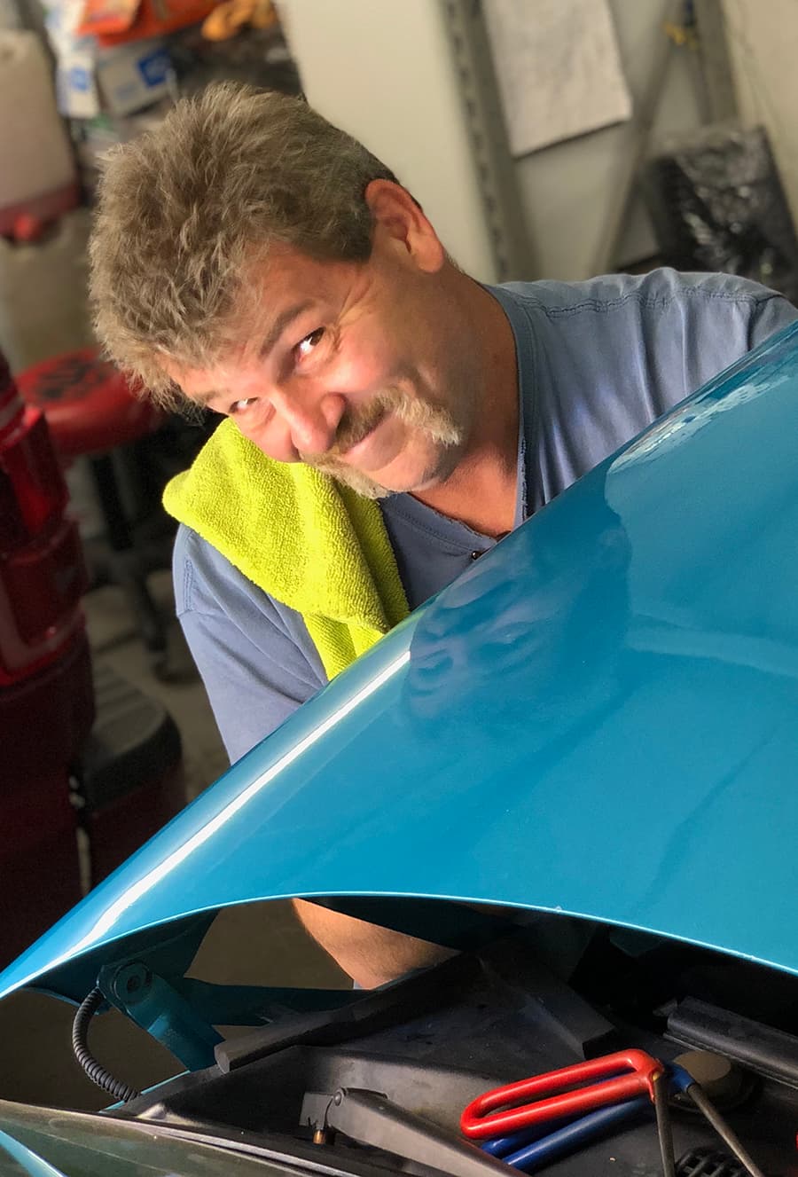 A Caropractor technician grinning over the hood of a teal car in the shop.
