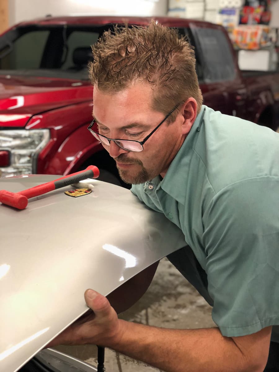 A Caropractor technician sights along a silver hood for dents, a red dent hammer resting on the panel.